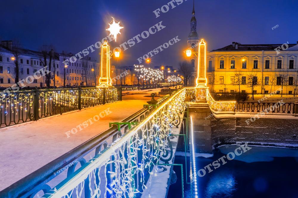 Фотография праздничного Санкт-Петербурга, Россия, сделанная в рождественский и новогодний вечер. На изображении показана улица с яркой праздничной иллюминацией и украшенный Старо-Никольский мост, перекинутый через Крюков канал. Этот фотоснимок передаёт атмосферу кануна Нового года: светящиеся гирлянды, отражения огней в воде и уютный зимний облик города. Фото подчёркивает красоту мостов Санкт-Петербурга и праздничное оформление улиц в период Рождества и Нового года в России. Иллюстрация может использоваться как изображение, картинка или слайд, посвящённые зимним праздникам и путешествиям по Санкт-Петербургу.